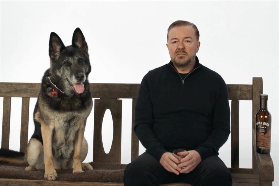 A dog and Ricky Gervaid sit on a bench, next to a bottle of Dutch Barn Vodka.