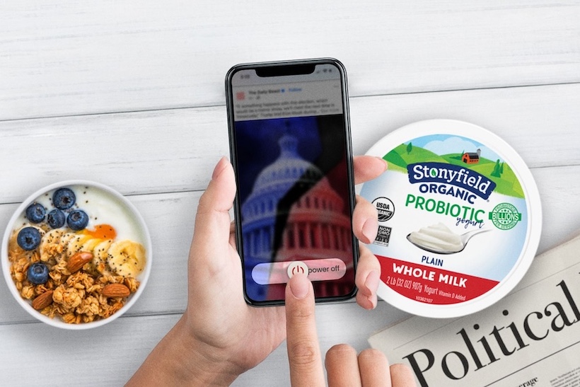 Hand holding smart phone displaying political content, with table set with Stonyfield products behind