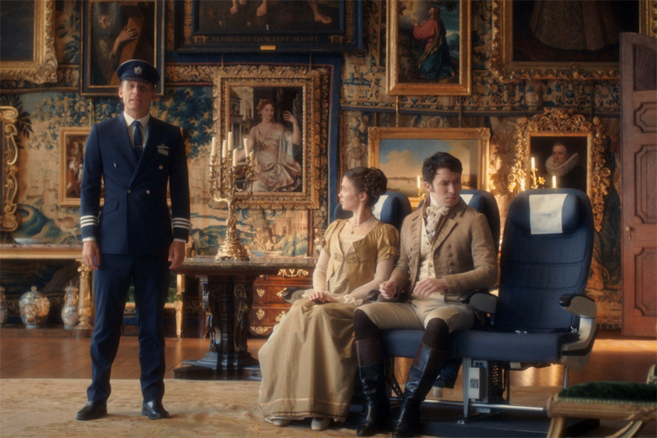 A BA pilot standing by two actors dressed in period drama costumes sitting on BA chairs